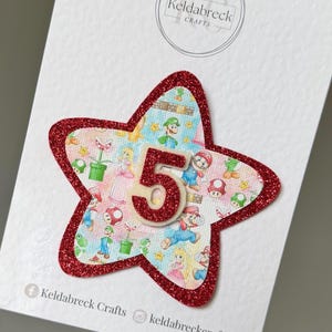 May include: A star-shaped decoration with a red glitter border and a colourful Super Mario Bros. design. The number "5" is in the centre. The text "Keldabreck Crafts" is at the bottom.