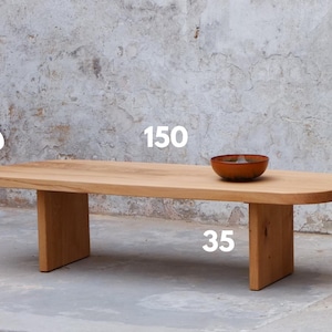 Può includere: Un tavolino da caffè in legno chiaro con piano ovale, lungo 150 cm, largo 50 cm e alto 35 cm. Una piccola ciotola rotonda marrone è appoggiata sul tavolo. Il tavolo ha due gambe rettangolari.