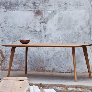 Op de afbeelding: Een rechthoekige houten eettafel met vier taps toelopende poten en een glad, vlak oppervlak. Een kleine, donkere, ronde kom staat aan de linkerkant van het tafelblad. De tafel is gemaakt van lichtgekleurd hout met een zichtbare houtnerf.