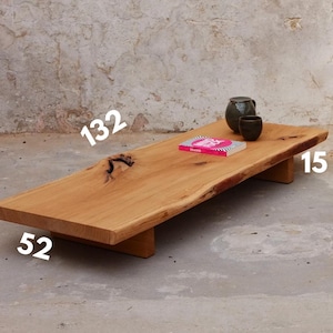 Puede incluir: Una mesa de centro de madera larga con un acabado natural y rústico. La mesa mide 132 cm de largo, 52 cm de ancho y 15 cm de alto. Tiene un diseño único de borde vivo y está soportada por dos patas de madera.