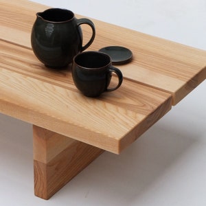 Könnte beinhalten: Ein heller Holz-Couchtisch mit einer rechteckigen Tischplatte und einem dreieckigen Sockel. Zwei dunkelgrüne Keramikbecher und eine Untertasse stehen auf dem Tisch.