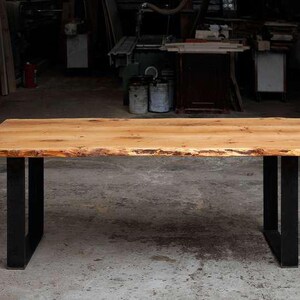Può includere: Un tavolo da pranzo rettangolare con un piano in legno marrone chiaro caratterizzato da un bordo naturale. Il tavolo è sostenuto da due robuste gambe in metallo nero a forma di U posizionate alle estremità, che offrono una base stabile.