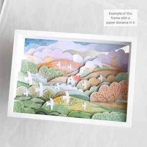 Może przedstawiać: Biała rama z papierowym diorama w środku. Diorama przedstawia krajobraz z wzg&oacute;rzami, drzewami, domem i latającymi ptakami. Niebo jest niebieskie z chmurami i gwiazdami. Wzg&oacute;rza są zielone, ż&oacute;łte i pomarańczowe. Drzewa są zielone i brązowe. Dom jest biały z czerwonym dachem. Ptaki są białe.