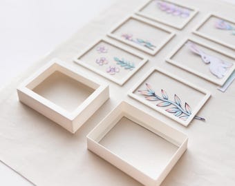 May include: A collection of rectangular white frames arranged on a surface. Some frames are empty, while others contain illustrations of a rabbit, floral patterns, and botanical sprigs in soft pastel colors, creating a layered visual effect.