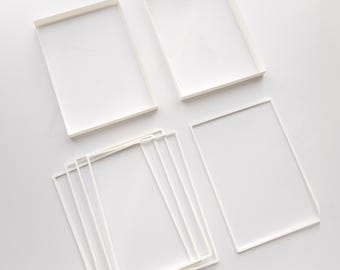 May include: A collection of white rectangular frames with thin borders. Two frames are positioned at the top, while a stack of four identical frames is arranged at the bottom left, and one single frame is placed on the bottom right side.