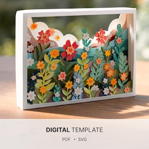 May include: A rectangular white shadow box frame containing a multi-layered paper craft scene of a colorful flower garden with rolling hills and clouds. The text DIGITAL TEMPLATE PDF SVG is printed below the frame on a plain background.