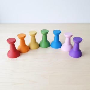 May include: A set of seven small, solid-coloured wooden pegs arranged in a semi-circle. The pegs feature a flared base and a rounded top, coloured in red, orange, yellow, green, blue, light pink, and purple, each with a smooth, matte finish.