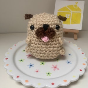 Könnte beinhalten: Ein gehäkeltes Mops-Spielzeug mit braunen Ohren, schwarzen Augen und einer herausgestreckten rosa Zunge. Das Spielzeug sitzt auf einem weißen Teller mit bunten Blumen.