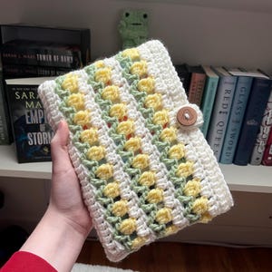 May include: A crocheted book cover with a white background and yellow and green stripes. The cover has a button closure.