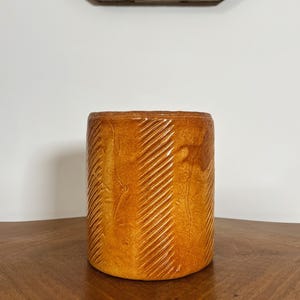 Peut inclure: Un récipient cylindrique en céramique émaillée de couleur ambre chaude. L'extérieur présente un motif diagonal répété de lignes gravées. Le récipient repose sur une surface en bois, avec un cadre de fenêtre blanc et vieilli en arrière-plan.