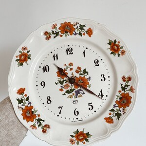 Könnte beinhalten: Ein weißer Keramikteller mit einem Blumendesign mit orangefarbenen und grünen Blumen. Der Teller hat ein Zifferblatt mit schwarzen Zahlen und Zeigern. Das Zifferblatt zeigt "Japy France".