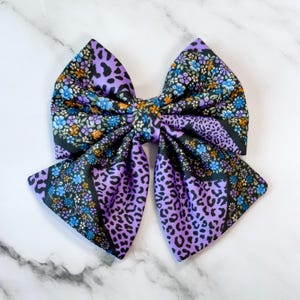 Può includere: Un grande fiocco decorativo per capelli con un mix di motivi. Il fiocco ha un nodo centrale e due grandi anelli. I motivi includono una stampa leopardata viola, una stampa floreale con fiori blu e arancioni e un bordo nero.