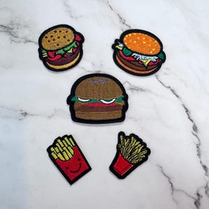 May include: Embroidered patches featuring burgers and french fries. The burger patches have brown buns, lettuce, tomato, and a beef patty. The french fry patches have red containers and yellow fries. The patches have black borders.