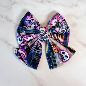 May include: A fabric hair bow featuring a vibrant, multi-colored print with purple, blue, and black tones. The pattern displays various cartoon characters, including a skeletal figure, a ghost, and a pumpkin, set against a starry night background.
