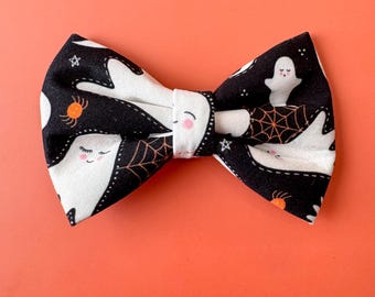 May include: A black bow tie featuring a repeating pattern of white ghosts with pink cheeks, orange spider webs, and small white stars. The fabric has a central knot detail and a structured shape, showcasing a festive Halloween-themed design.