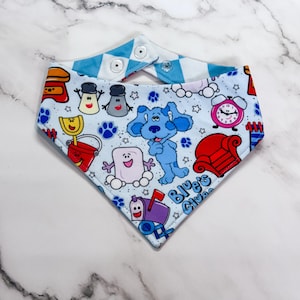 May include: A triangular bandana featuring a light blue background with a repeating pattern of Blue's Clues characters and objects, including Blue the puppy, a red chair, a mailbox, and a pink alarm clock, with two white snap buttons at the top.