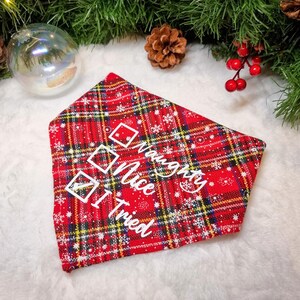 May include: Red plaid pet bandana with white text that reads "Naughty, Nice, I Tried" and check boxes. The bandana has a festive holiday theme with snowflakes and a Christmas tree in the background.