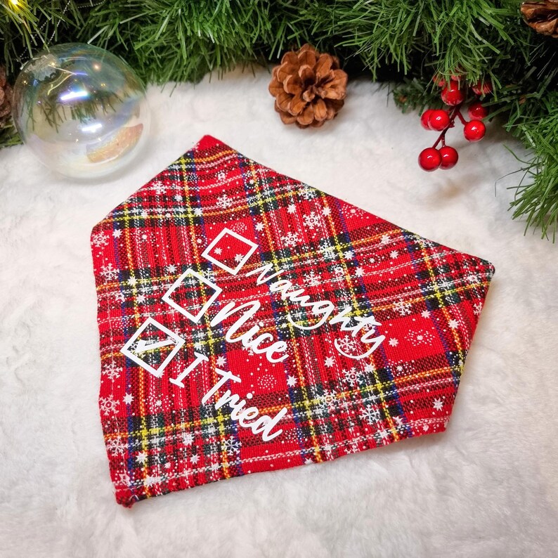 May include: Red plaid pet bandana with white text that reads "Naughty, Nice, I Tried" and check boxes. The bandana has a festive holiday theme with snowflakes and a Christmas tree in the background.