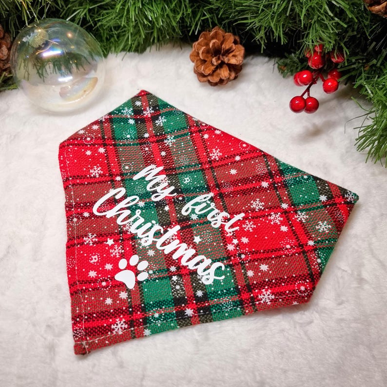 May include: A red, green, and brown plaid dog bandana with the text "My first Christmas" and a white paw print. The bandana is decorated with white snowflakes. Festive pet accessory for the holiday season.