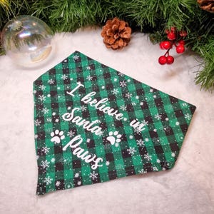 May include: A green and black plaid dog bandana with white text that reads "I believe in Santa Paws" and paw prints. The bandana has a snowflake pattern and is surrounded by Christmas decorations.