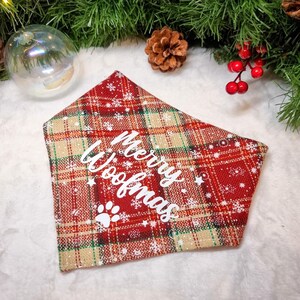 May include: Red plaid dog bandana with white text reading "Merry Woofmas" and a paw print. The bandana has a festive design with white snowflakes and stars. The background includes Christmas decorations.