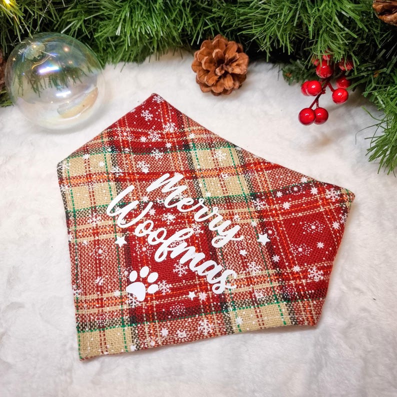 May include: Red plaid dog bandana with white text reading "Merry Woofmas" and a paw print. The bandana has a festive design with white snowflakes and stars. The background includes Christmas decorations.