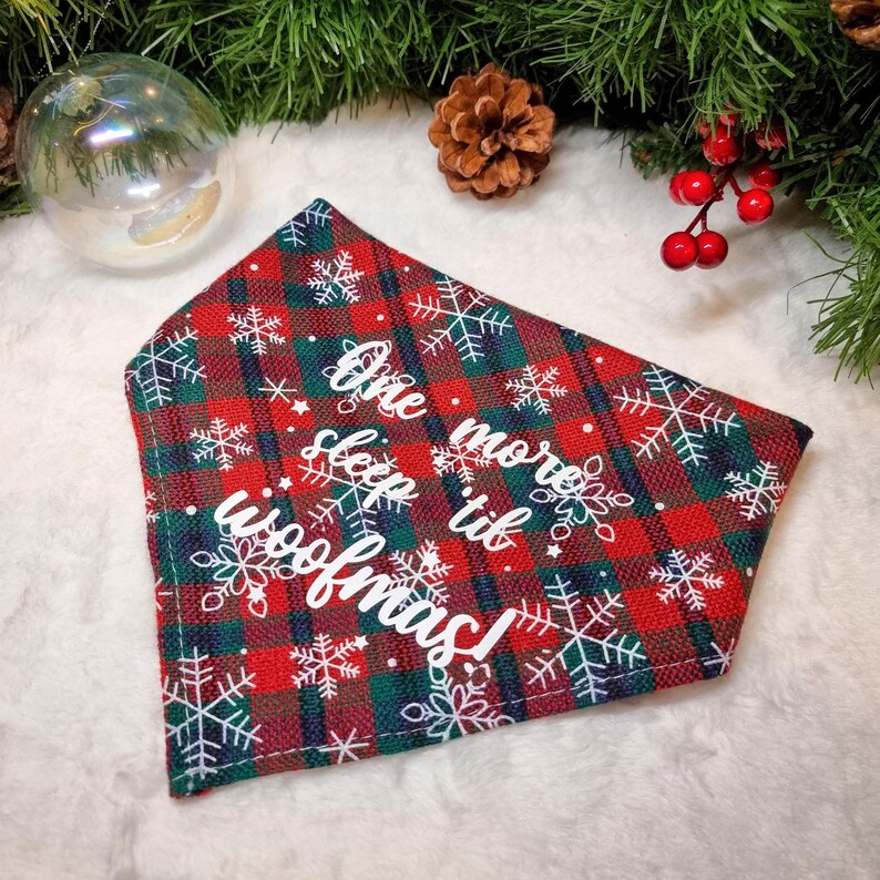 May include: A red, green, and white plaid dog bandana with white snowflakes and the text "One more sleep til woofmas!". The bandana is decorated with Christmas-themed elements, including a pine cone and red berries.