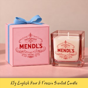 May include: A square pink gift box with a light blue ribbon and a white scented candle in a square pink glass container. Both the box and the candle feature red text that reads Patisserie Mendl's, Brucknerplatz, No.12, Nebelsbad.