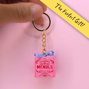 Può includere: Un portachiavi in metallo dorato con un ciondolo quadrato rosa sormontato da un fiocco in nastro azzurro. Il ciondolo presenta la scritta rossa in rilievo PATISSERIE MENDL'S, insieme a decorazioni e alla scritta Nebelsbad.