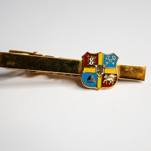 Puede incluir: Un pasador de corbata dorado con un escudo de esmalte de colores que presenta un escudo con un número 8, una llave, un pájaro y un caballo.