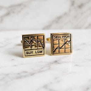 May include: A pair of square gold-toned metal cufflinks. One cufflink features a grid pattern with a downward trending line and the text BUY LOW. The other cufflink features a grid pattern with an upward trending line and the text SELL HIGH.
