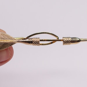 May include: A gold-toned tie tack in the shape of a dart. The dart has a gold-toned fletching and a gold-toned tip.