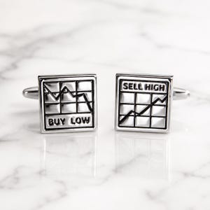 May include: A pair of square silver-toned cufflinks featuring a grid pattern and black engraved text. One cufflink displays a downward trend line with the text BUY LOW, while the other shows an upward trend line with the text SELL HIGH.