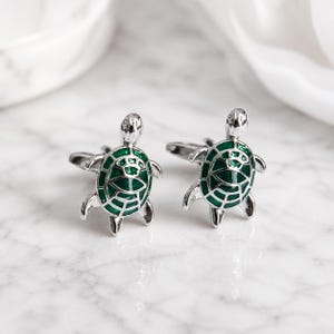 May include: A pair of silver-toned cufflinks shaped like sea turtles. Each turtle features a polished metallic finish with a vibrant green enamel inlay on the shell, highlighting the detailed geometric pattern of the carapace and flippers.