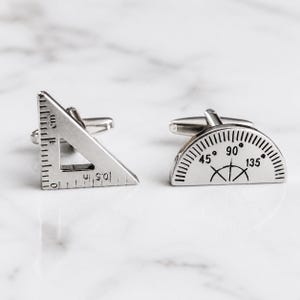 May include: A pair of silver-toned metal cufflinks. One cufflink is shaped like a right-angle triangle ruler with markings for 1 cm and 0.5 in. The other cufflink is shaped like a semi-circular protractor with degree markings for 45, 90, and 135.