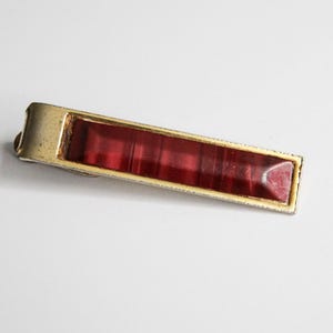 May include: A vintage tie clip featuring a rectangular, deep red, textured insert set within a gold-toned metal frame. The clip is designed to secure a tie and has a classic design.