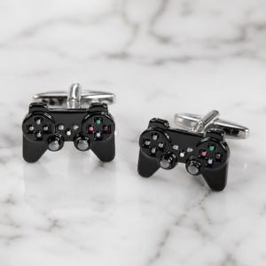 May include: A pair of black cufflinks shaped like video game controllers. Each cufflink features a glossy black finish with silver-toned metal hardware on the back, including a small toggle bar. The controller design includes small colored buttons.