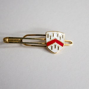 Puede incluir: Un broche de corbata de oro con un diseño de escudo de esmalte blanco y rojo. El escudo tiene un galón rojo y múltiples flechas doradas.