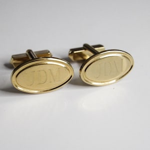 Op de afbeelding: Een paar goudkleurige manchetknopen met een ovaal ontwerp. Elke manchetknop heeft de gegraveerde initialen "JDM". De manchetknopen zijn geplaatst tegen een witte achtergrond.
