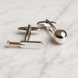 May include: A pair of silver-toned cufflinks featuring a cricket bat design with gold-toned detailing on the handle and a spherical cricket ball design with gold-toned stitching accents. The cufflinks have a polished metallic finish throughout.