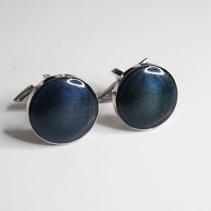 Può includere: Un paio di gemelli rotondi con una superficie blu scuro a motivo a griglia. I gemelli hanno un bordo in metallo color argento e un retro a cerniera. Lo sfondo è una superficie bianca.