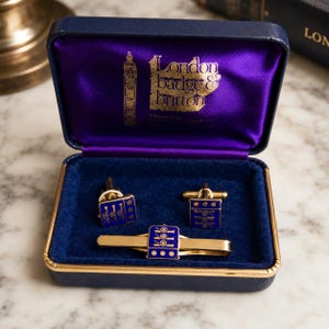 May include: A gold-tone tie bar and a pair of matching cufflinks featuring a blue enamel shield with three gold cannons and three dots. The set is presented in a dark blue hinged box with a purple satin lining and the text London Badge & Button.