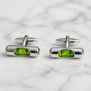 Può includere: Un paio di gemelli in metallo color argento con un design a livella a bolla cilindrica. Ogni gemello contiene una fiala di liquido verde brillante con una bolla d'aria centrata, racchiusa in una cornice di metallo lucido con chiusura a perno.