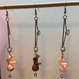 Op de afbeelding: Drie paar oorbellen met een zilveren ketting en een klein plastic figuurtje dat eronder hangt. De figuurtjes zijn naakt, bruin en roze.