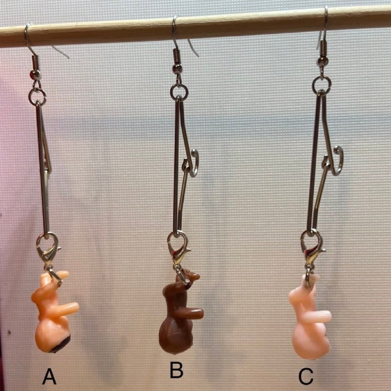 Op de afbeelding: Drie paar oorbellen met een zilveren ketting en een klein plastic figuurtje dat eronder hangt. De figuurtjes zijn naakt, bruin en roze.