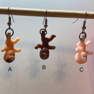 Op de afbeelding: Drie paar hangende oorbellen met omgekeerde babypoppen. De poppen zijn gemaakt van plastic en zijn verkrijgbaar in drie huidtinten: lichtroze, bruin en perzik.