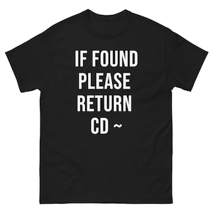 Peut inclure: T-shirt noir avec l'inscription blanche "IF FOUND PLEASE RETURN CD ~" imprimée sur le devant. Le t-shirt a des manches courtes et un col rond. Le texte est en gras, sans empattement.