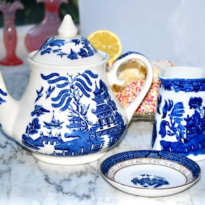 Puede incluir: Una tetera, una jarra de leche y un platillo de porcelana azul y blanco con un patrón tradicional de sauce. La tetera tiene una tapa y un asa. La jarra de leche tiene un asa y un pico. El platillo tiene un borde dorado.