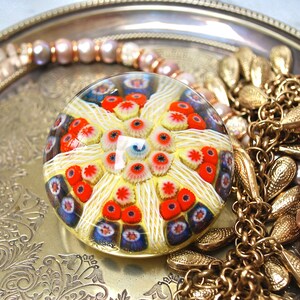 May include: A colorful glass paperweight with a white, red, and blue pattern. The paperweight is round and has a clear glass exterior. It is sitting on a silver tray with gold jewelry.