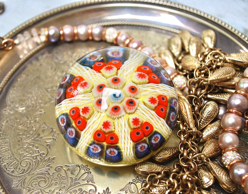 May include: A colorful glass paperweight with a white, red, and blue pattern. The paperweight is round and has a clear glass exterior. It is sitting on a silver tray with gold jewelry.